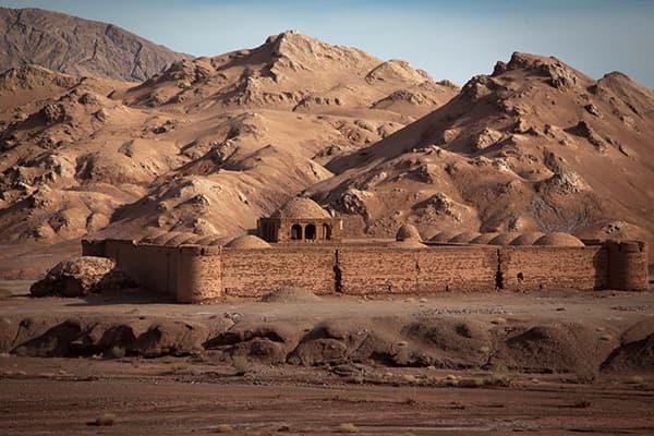 Historic caravanserai in the Kavir Desert of Iran - @advandortmontduurzaam 04/2025 Historic brick caravanserai in the Kavir Desert of Iran, framed by rugged, reddish-brown desert mountains under a clear blue sky. The rectangular fortress-like structure features domed roofs, arched entrances, and watchtower corners, once serving as a safe resting place for travelers and traders along ancient desert trade routes. The barren, dry landscape surrounding the caravanserai highlights the isolation and stark beauty of the Iranian desert, an important stop on Ad van Dortmont’s overland expedition from the Netherlands to Pakistan.