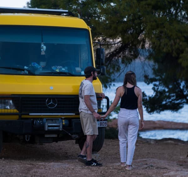 Astrid & Jaron with their Mercedes Camper "Morris" - @our_orbit_vanlife 07/2024 A couple, @our_orbit_vanlife, stands hand in hand beside their yellow Mercedes Vario 816D camper van parked in a scenic outdoor setting with trees and a body of water in the background.