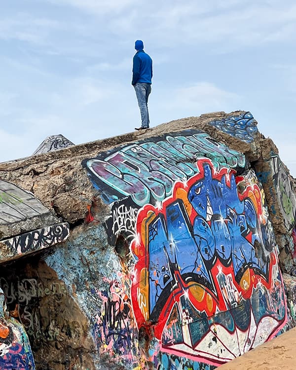 Standing atop the remains of a WWII-era bunker on the west coast of France - @van4life__ 04/2025 Chris from @van4life__ stands atop a large, graffiti-covered World War II-era bunker on the beach in Capbreton, France. Wearing a blue hoodie and jeans, he looks out over the horizon under a pale sky, capturing a moment of reflection during his vanlife journey. The concrete structure is vividly painted with colorful street art, contrasting sharply with the natural surroundings of sand and sea.