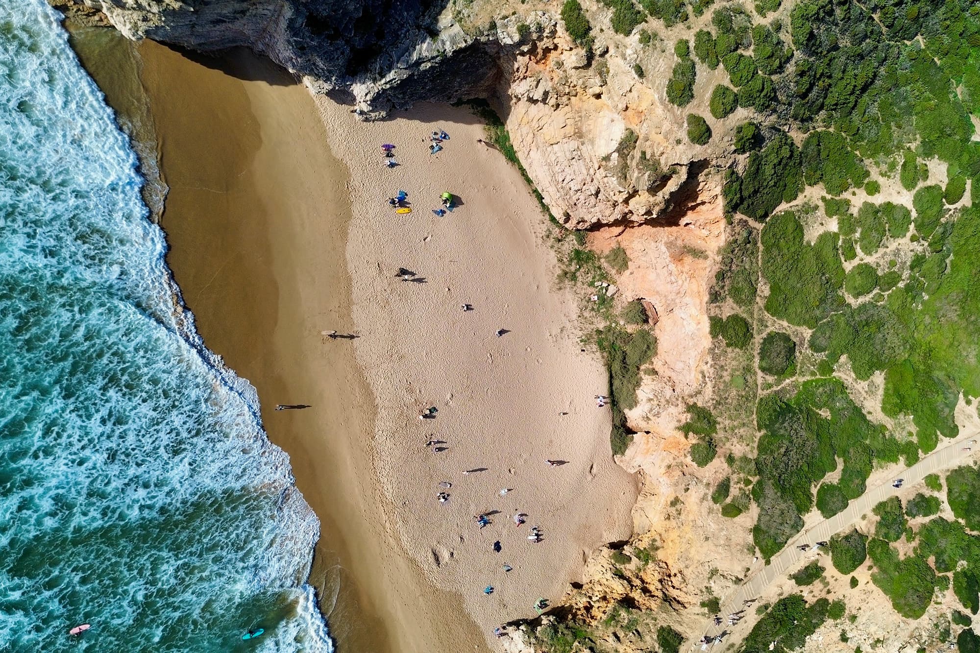 A drone view of one of Portugal's surf beaches - @van4life__ 04/2025 Drone view captured by @van4life__ of a hidden beach near Sagres, Portugal, where turquoise Atlantic waves crash onto golden sand. The scene is framed by dramatic cliffs and lush green hills. Surfers and sunseekers dot the shore, perfectly capturing the carefree, adventurous spirit of vanlife along the Iberian Peninsula.