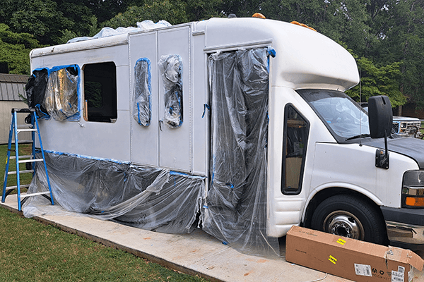 Renovating Lupine - driftwoodartwork 06/2025 A white Chevrolet E3500 shuttle bus is shown mid-renovation in a grassy backyard, with plastic sheeting and painter’s tape covering the windows and lower panels. A blue ladder stands beside the bus, and a cardboard box lies near the front tire. Artist Allison Leah Thompson, known on Instagram as @idonthavegum, is converting the vehicle into a mobile art studio and home for her solo travels as a nomadic artist and Black female traveler.
