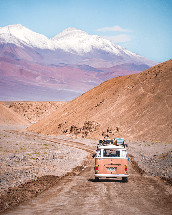 Exploring the Atacama region with my VW Kombi - @octaviaviajando 09/2022 Victoria, the Chilean traveler behind @octaviaviajando, drives her pink and white 1985 Volkswagen T2 Kombi down a winding dirt road in the Atacama Desert. The campervan is fully loaded with gear on the roof as it heads toward the snow-capped peaks of the Andes under a clear blue sky. This scene captures her solo vanlife journey through northern Chile, highlighting remote adventure, freedom, and sustainable overland travel through one of the most stunning desert landscapes on Earth.